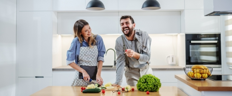 a man and a woman prepare vegetables and laugh at a kitchen island